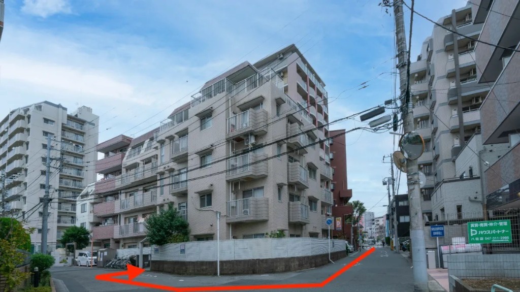マンション外観。赤い矢印の通り、建物手前のエントランス方面へ